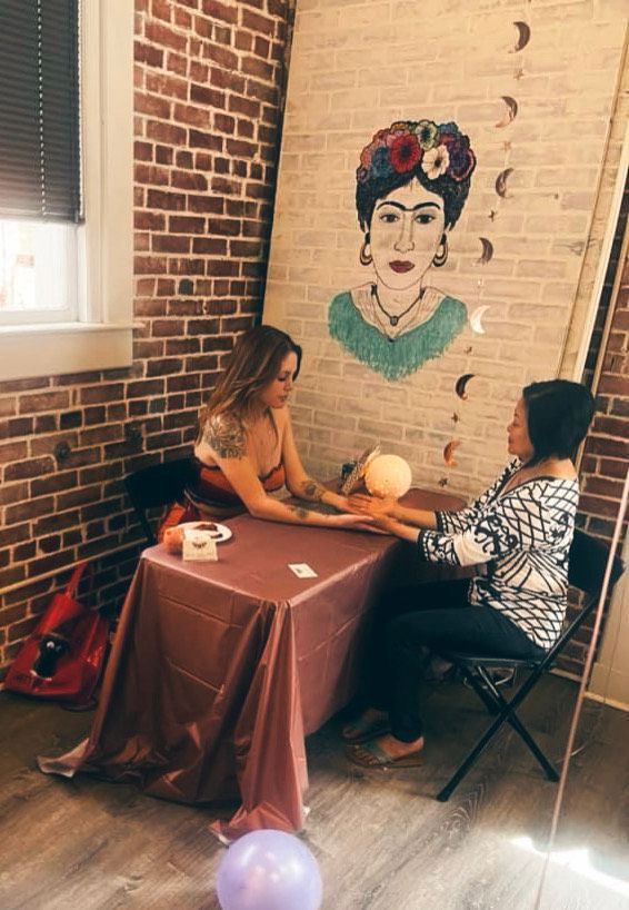 A woman is sitting at a table with a painting of frida kahlo on the wall.