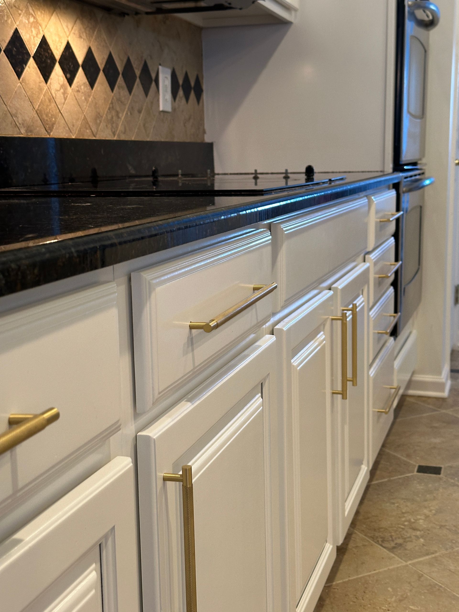 White kitchen cabinets with gold handles, black countertop, and diamond-patterned backsplash.