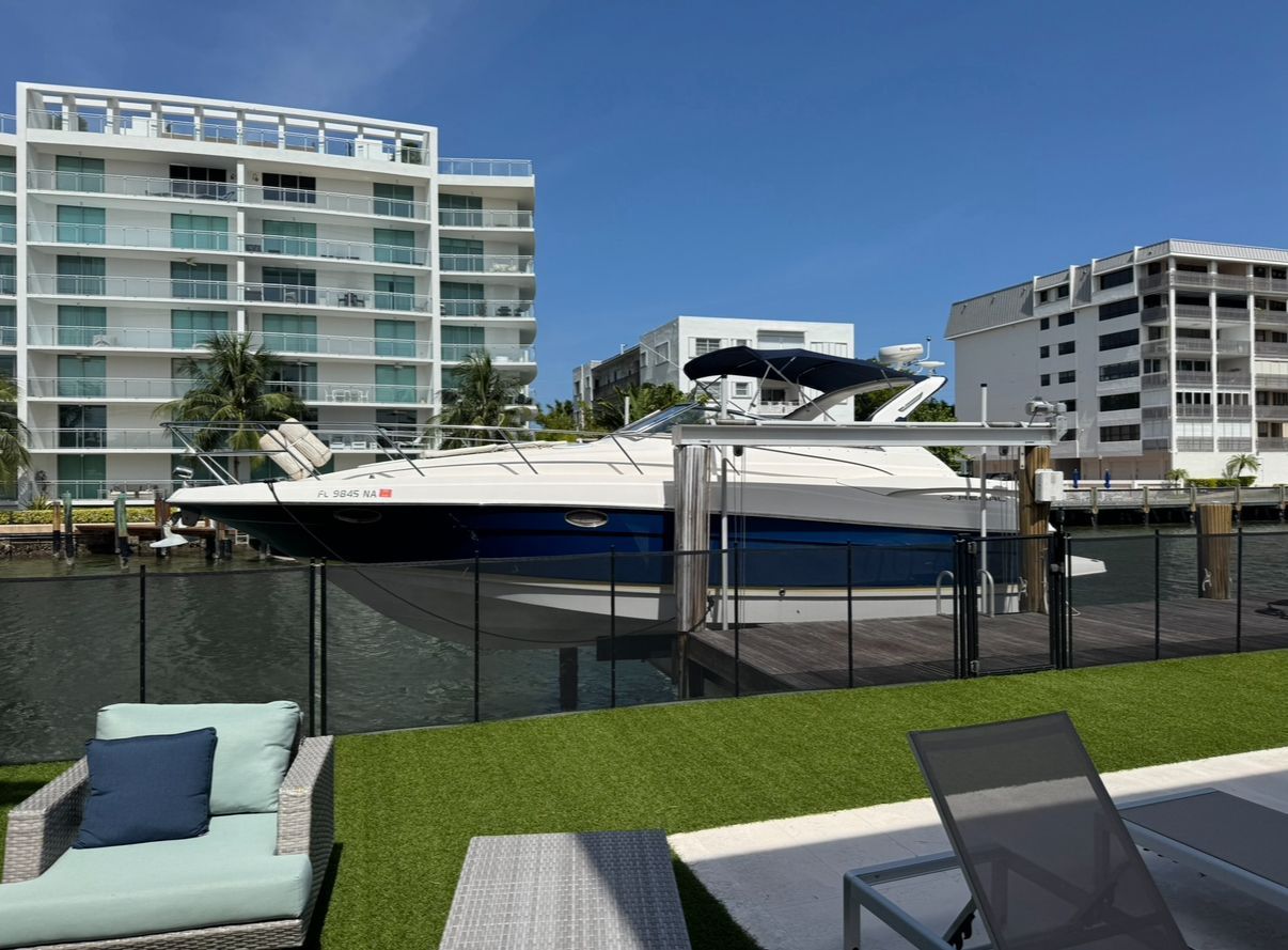 A yacht docked in a canal, flanked by buildings, with lounge chairs on a lawn in the foreground.