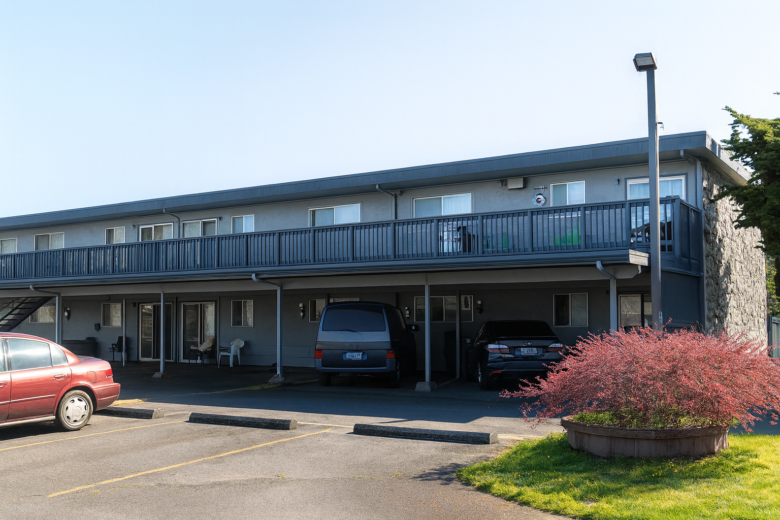 Two-story apartment building with parking, a second-floor balcony, and a bush with red leaves.