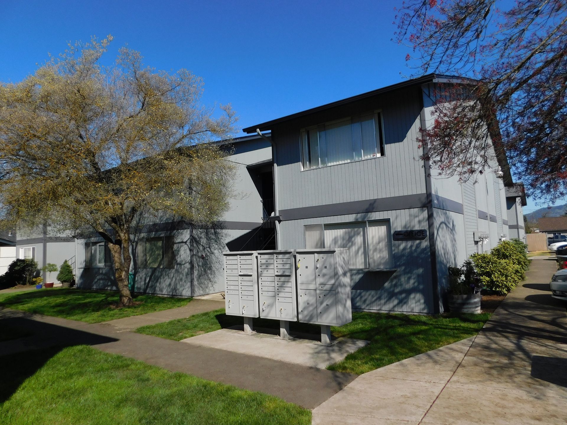 Gray apartment building with mailboxes, grass, and a clear blue sky.