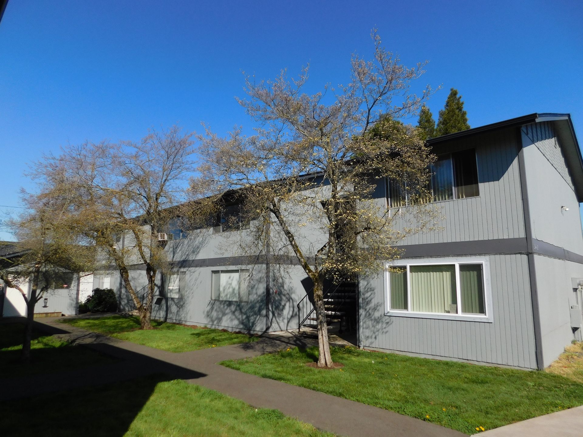 Two-story gray apartment building with trees and walkway on a sunny day.