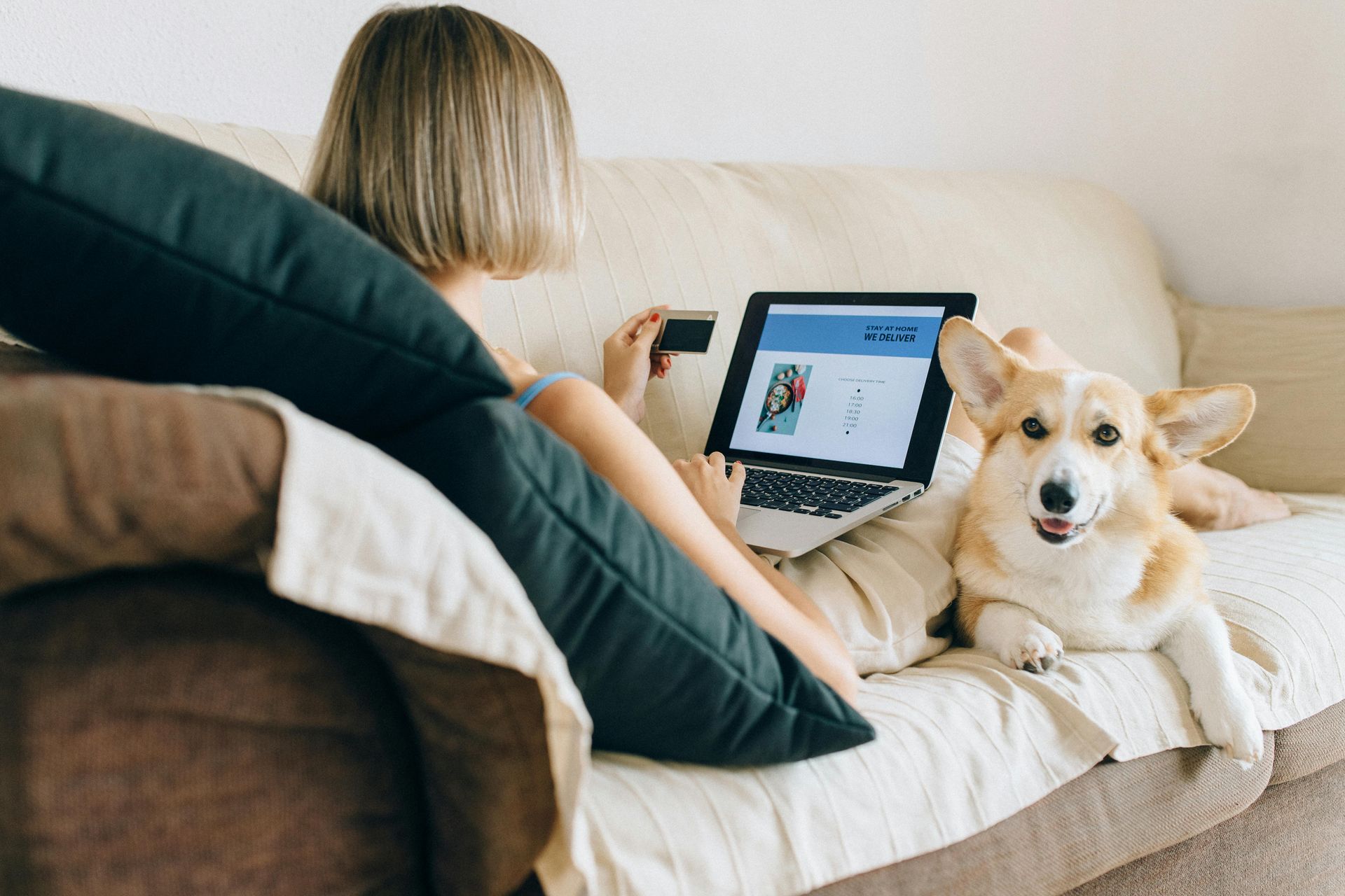 Woman using laptop on couch, dog lying next to her, both indoors.