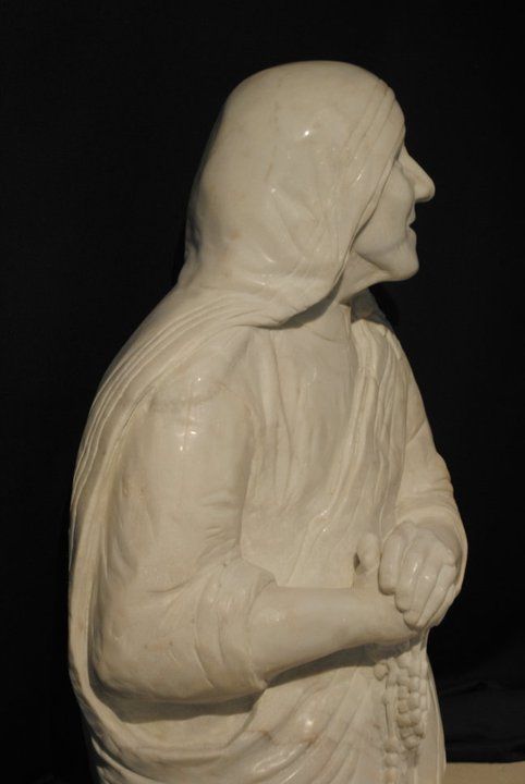 Estatua de mármol blanco de la Madre Teresa, de perfil, con las manos juntas y llevando un velo.