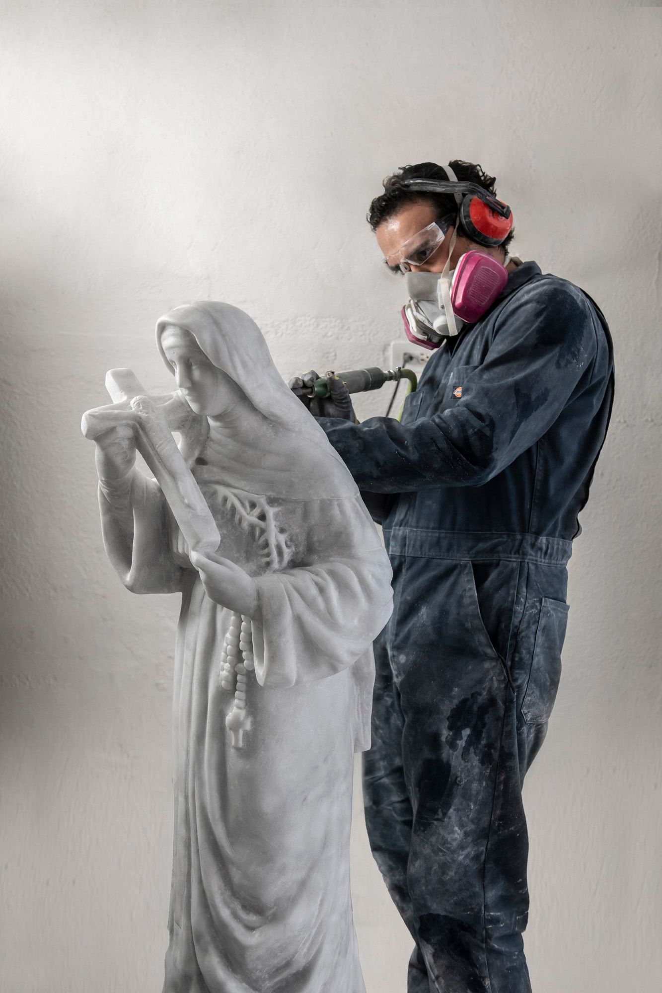 Un escultor vestido con un mono azul utiliza una herramienta eléctrica sobre una estatua de mármol blanco; lleva gafas de seguridad, máscara y protección para los oídos.