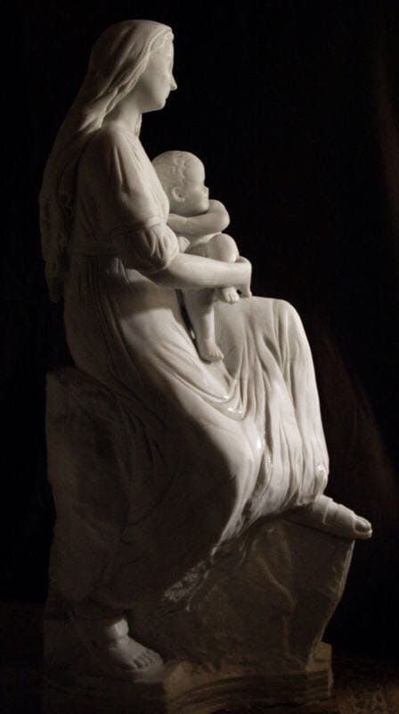 Escultura de mármol de una mujer sentada con un niño en brazos, de perfil, con ropa drapeada. Fondo oscuro.