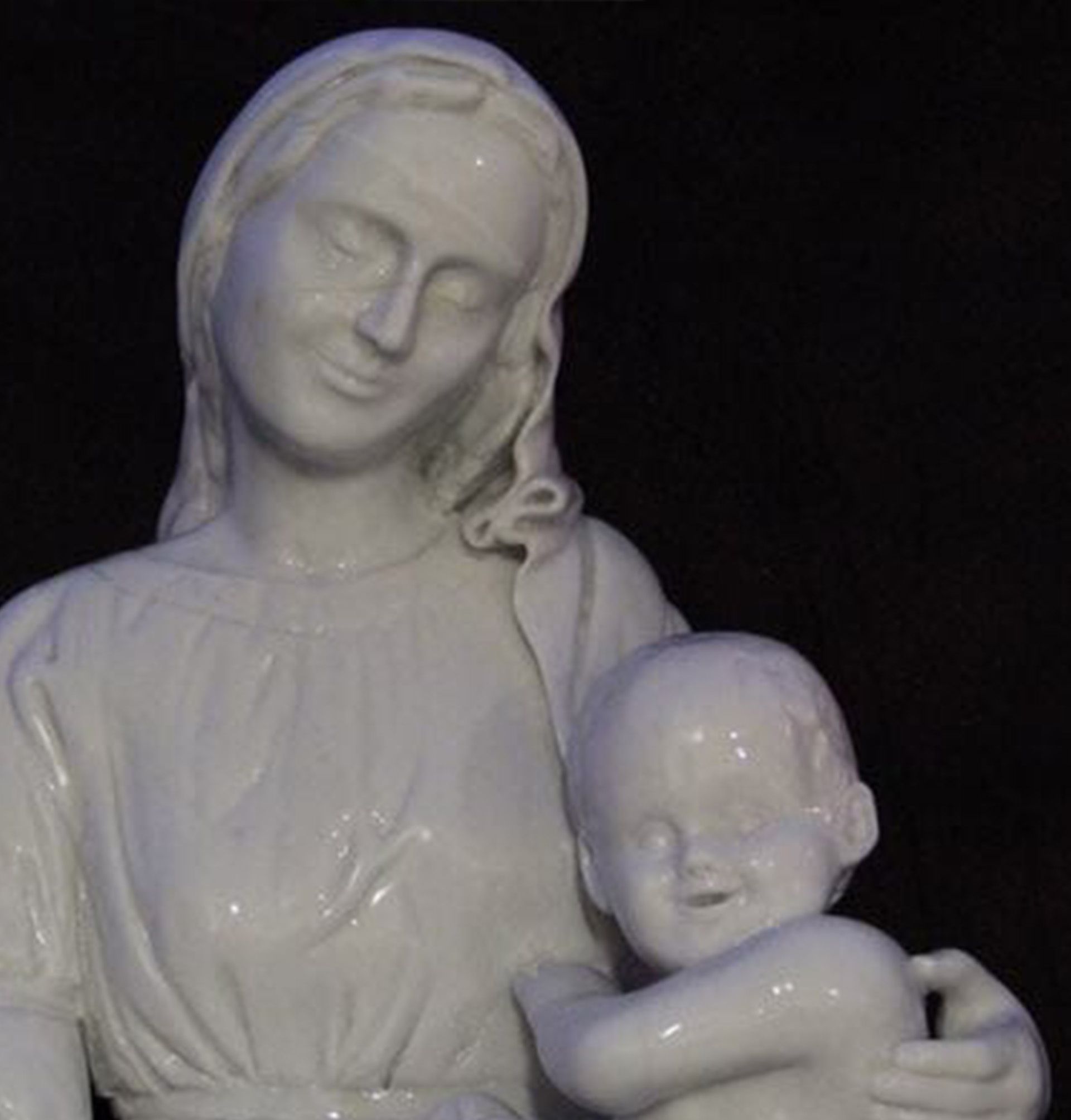 Estatua de cerámica blanca de María y el Niño Jesús, ambos con los ojos cerrados, sobre un fondo oscuro.