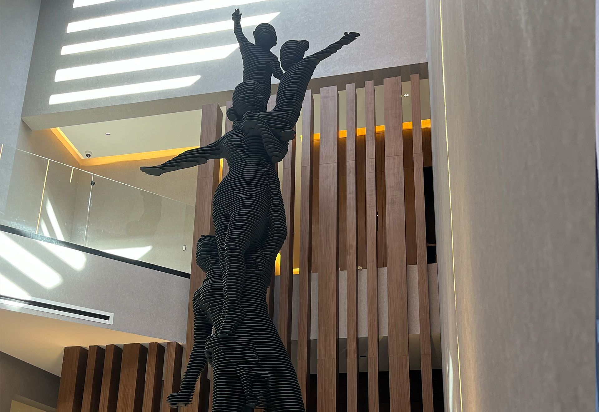 Escultura de tres figuras que se elevan hacia arriba, en un vestíbulo con paneles de madera y ventanas.