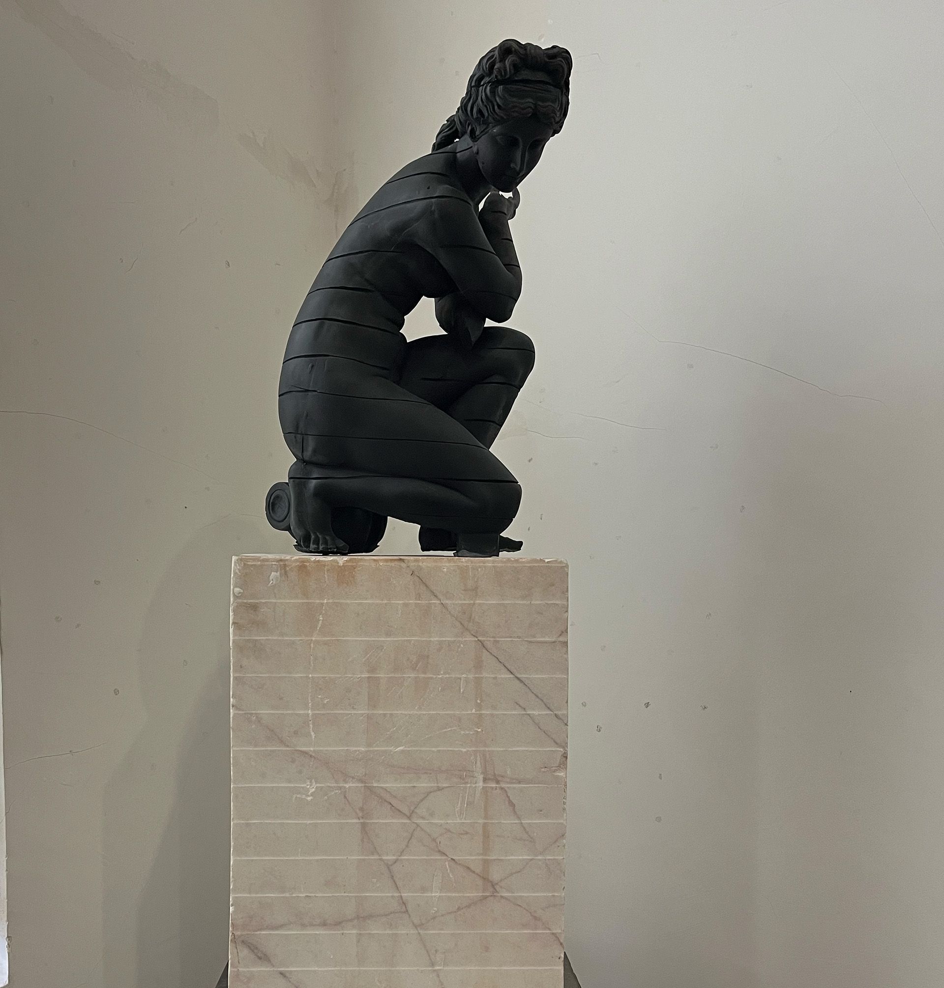 Escultura negra de una mujer arrodillada sobre un pedestal de mármol, en una habitación.