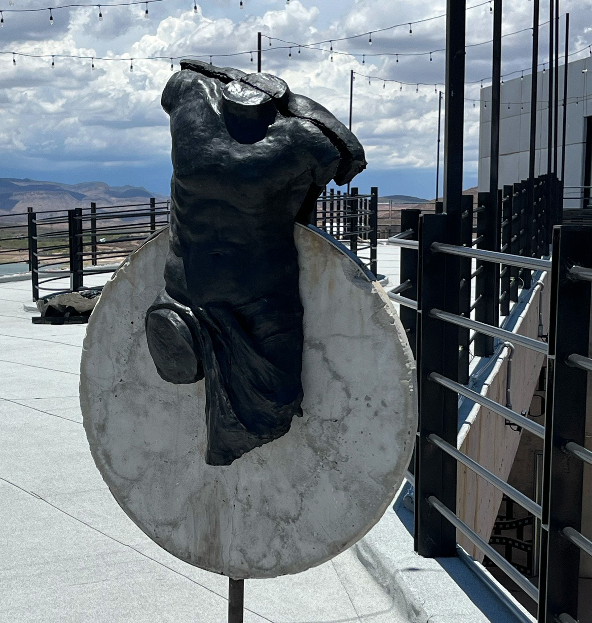 Estatua de torso negro sobre un disco de hormigón; entorno de azotea con cielo nublado y barandilla de metal.