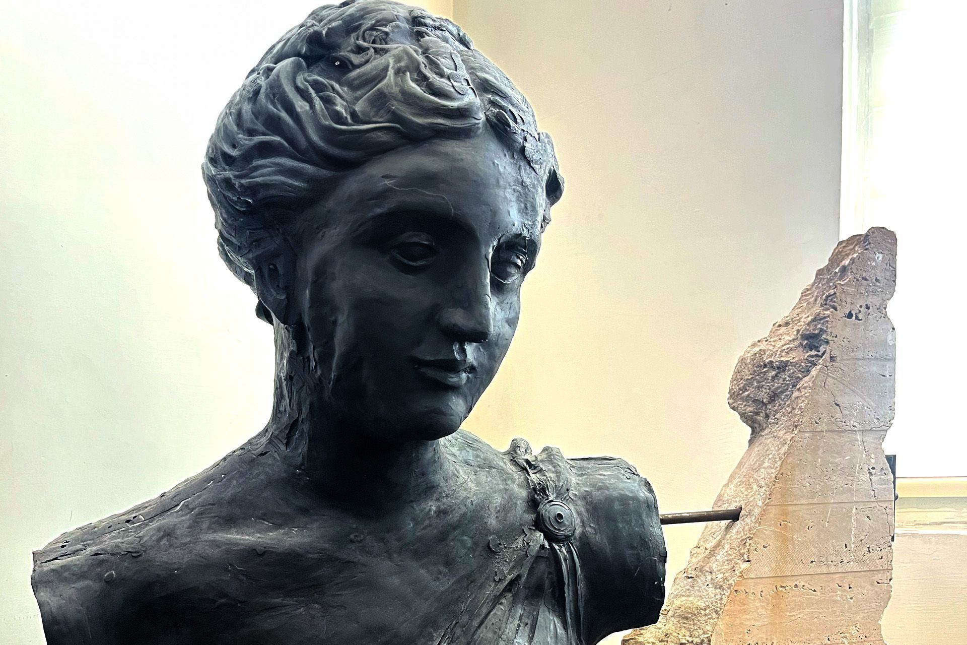 Un busto oscuro de mujer con rasgos clásicos, junto a un fragmento de piedra, en una habitación iluminada.