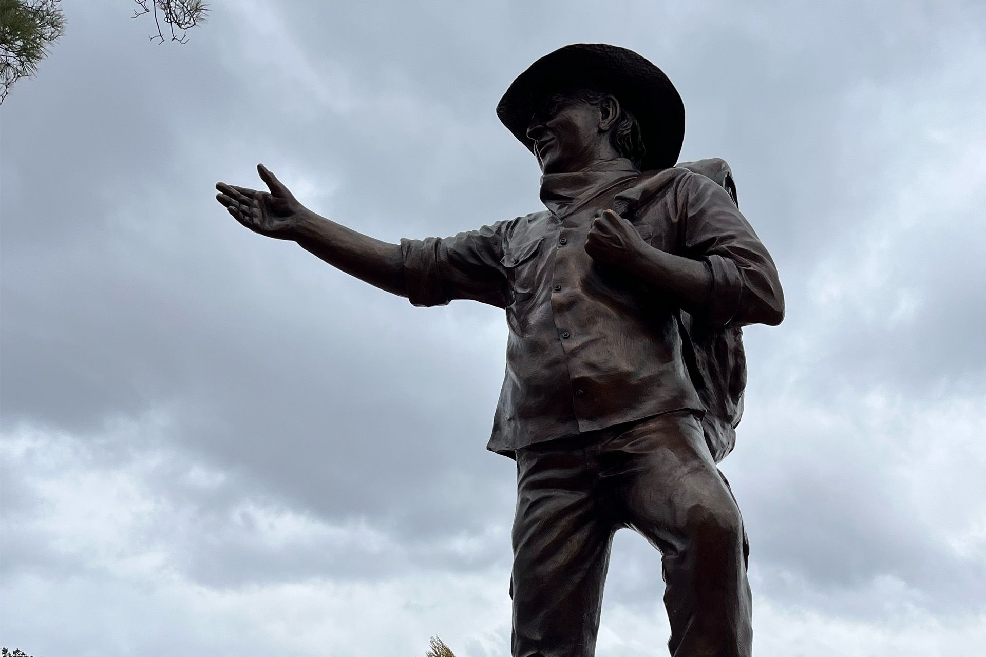 Estatua de bronce de un hombre con sombrero, brazo extendido y llevando una bolsa, frente a un cielo nublado.