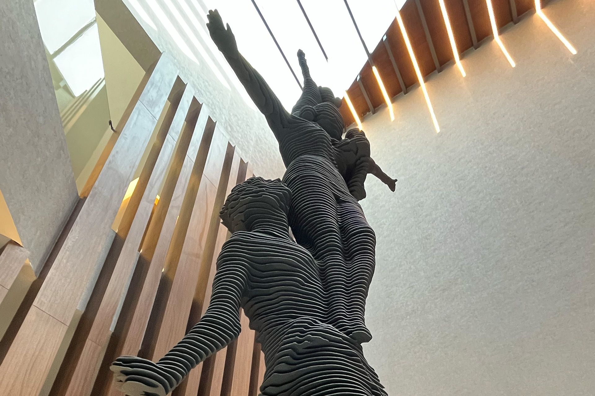 Escultura de figuras de acero que se extienden hacia arriba, en un entorno de edificio moderno con acentos de madera y luz natural.