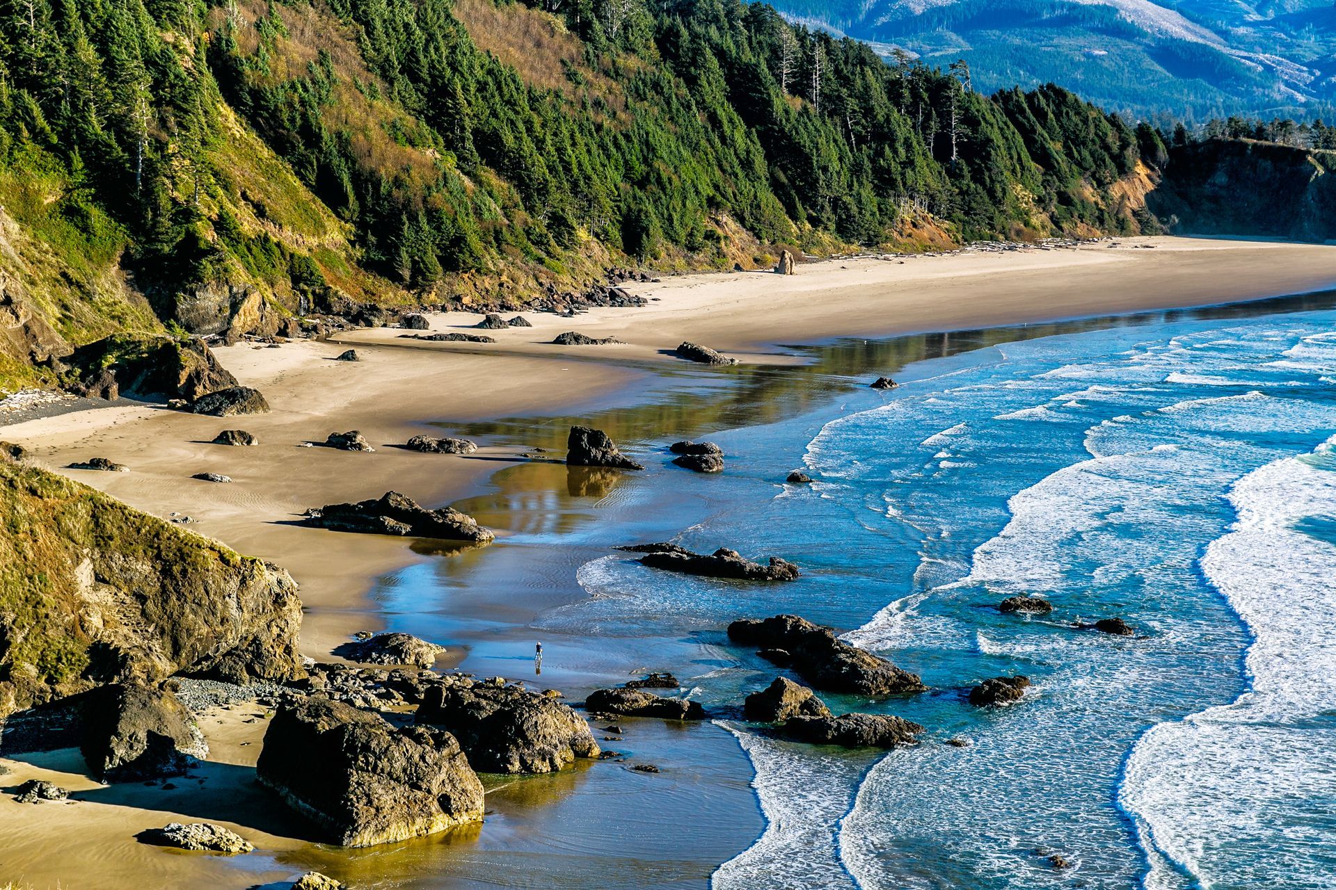 Sandy beach with rocky coast, blue ocean, green cliffside, and mountains in the distance.