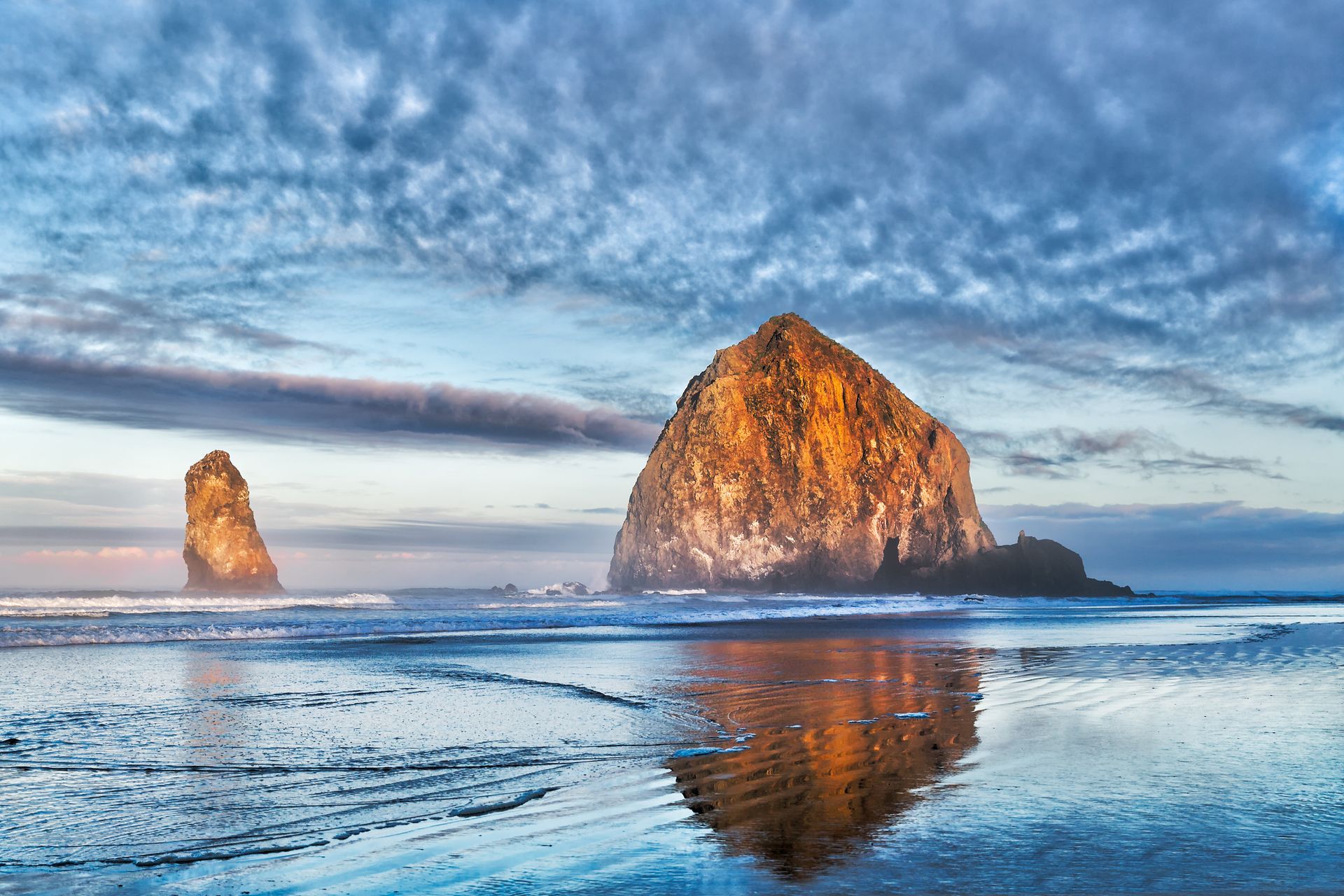 Two large sea stacks on a beach reflect in wet sand under a cloudy blue sky.
