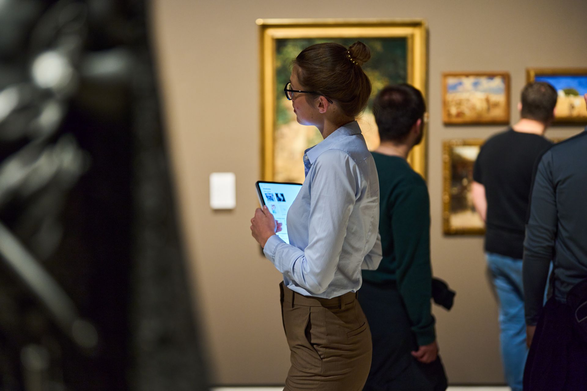 Woman in a museum looking at paintings, holding a tablet. Other visitors are nearby.