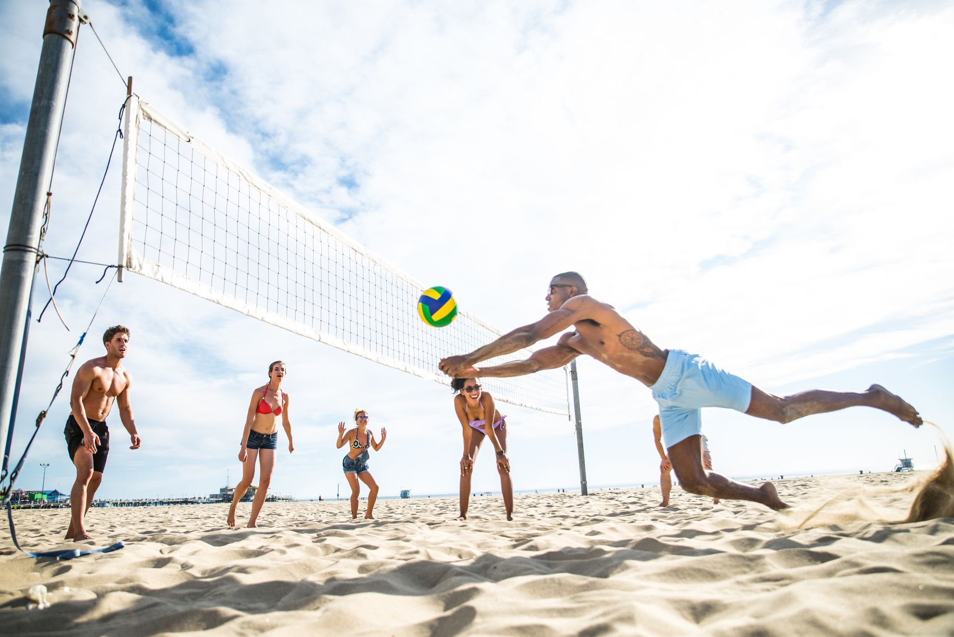 People playing beach volleyball on a sunny day. A man dives to hit the ball, others watch.