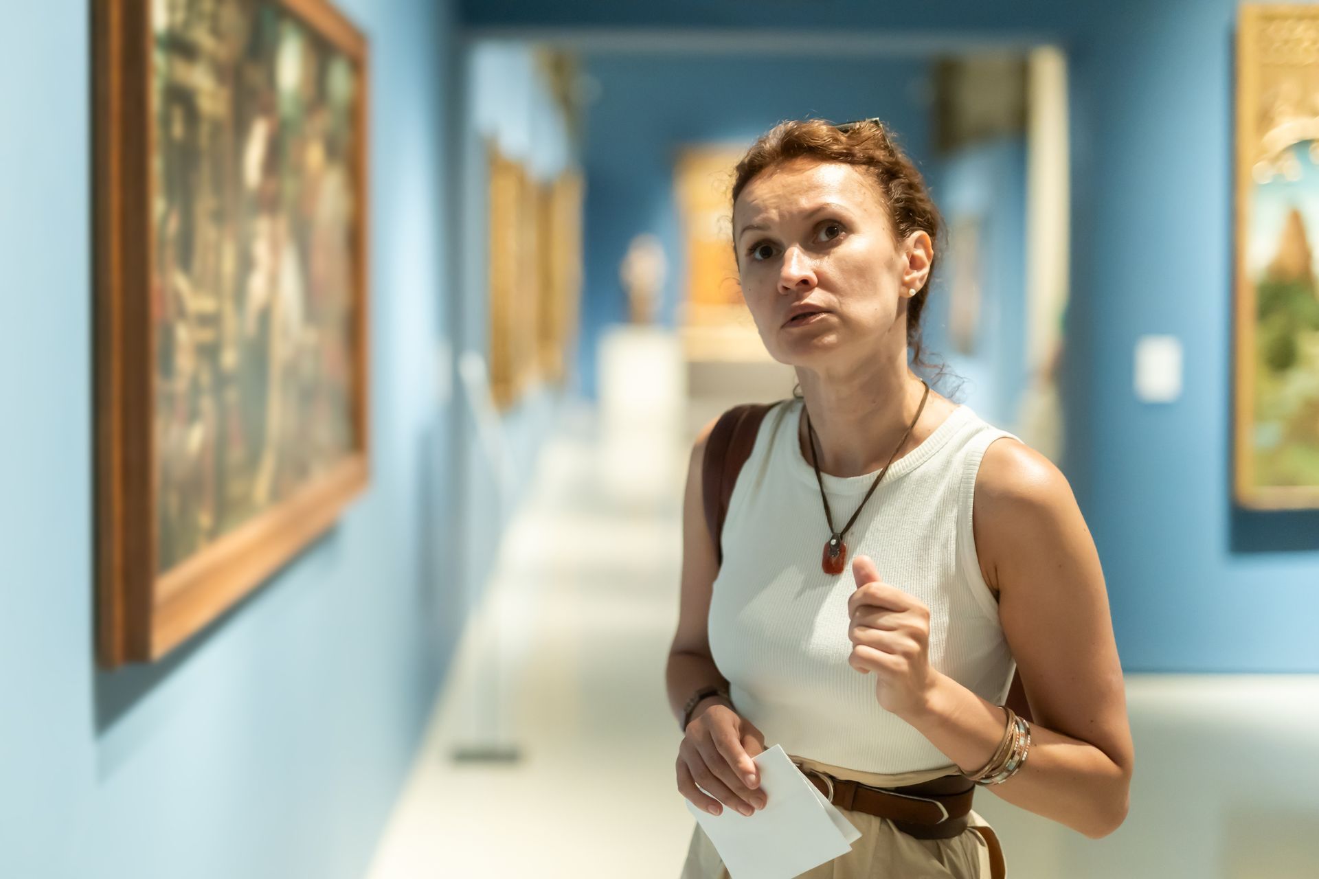 Woman in a white top and tan pants looks at paintings in a museum, pale blue walls.