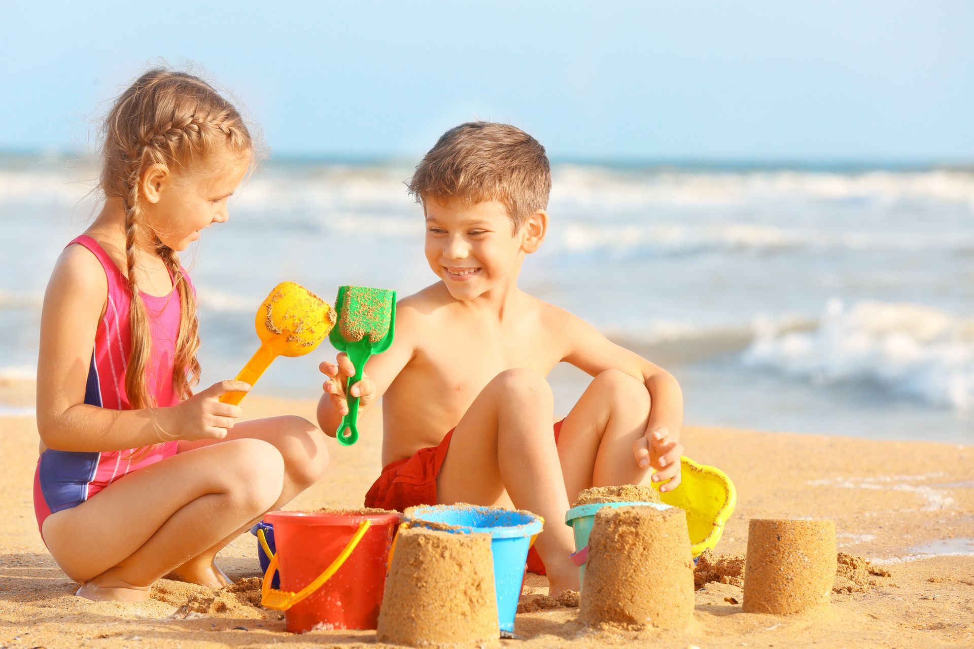 Girl and boy building sandcastles on a beach, smiling with sand toys in hand.