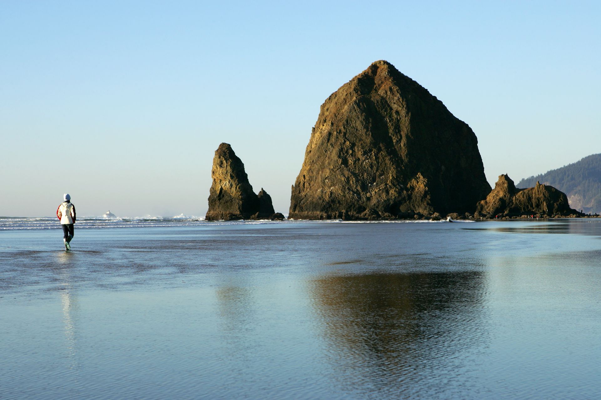 Person walking on wet sand toward large sea stack, Cannon Beach, Oregon, on a sunny day.