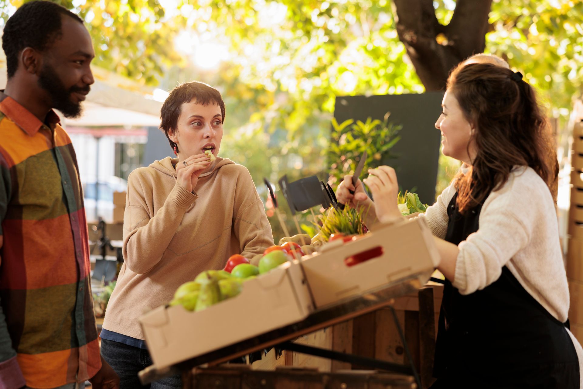 People at farmers market; woman eating sample, vendor offering produce.