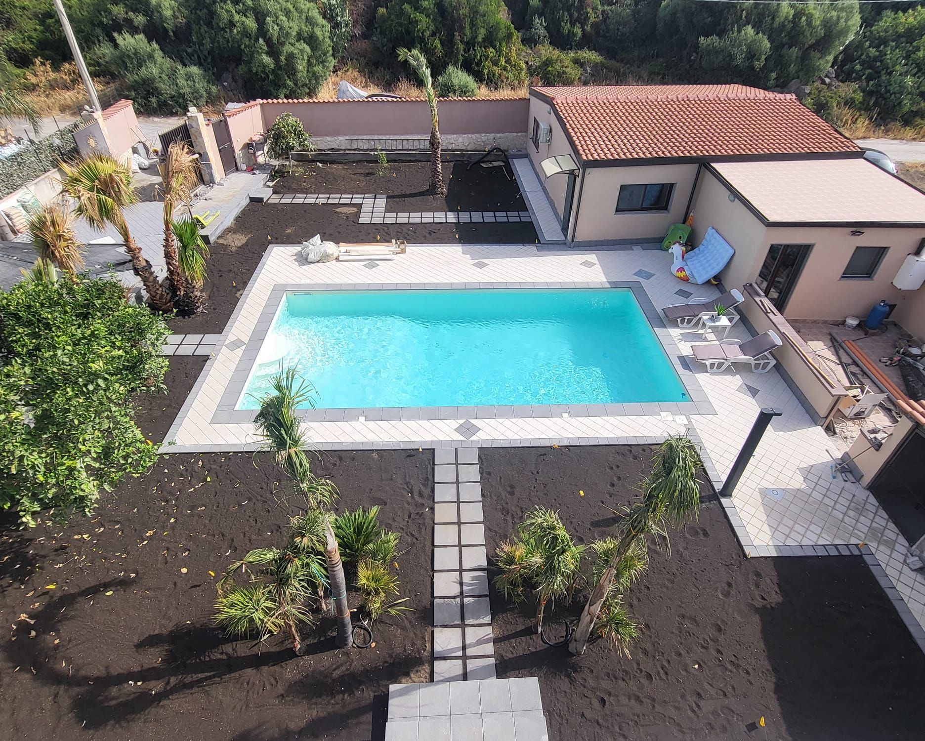 Vista aerea di una piscina rettangolare circondata da terreno scuro e da un patio pavimentato, vicino a una piccola casa.