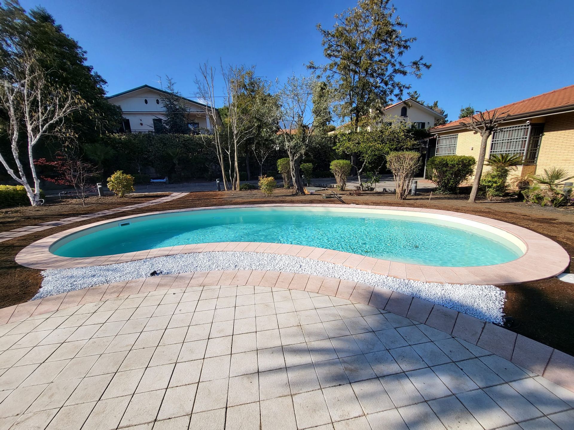Piscina di forma ovale circondata da un patio in mattoni, ciottoli bianchi ed erba. L'acqua azzurra riflette un cielo limpido e soleggiato.