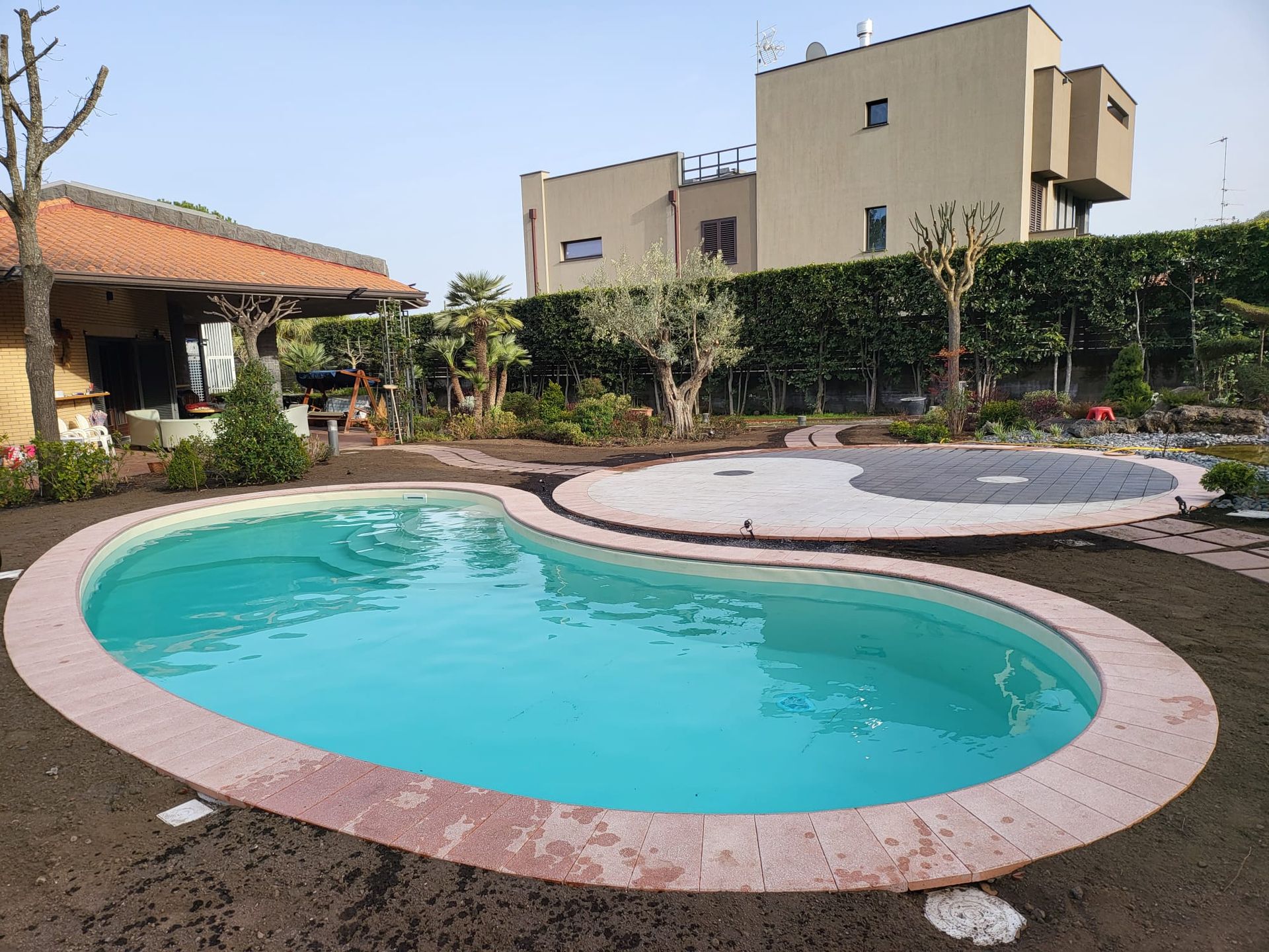 Piscina in un cortile con un design yin e yang, circondata da un giardino, un edificio e un gazebo.
