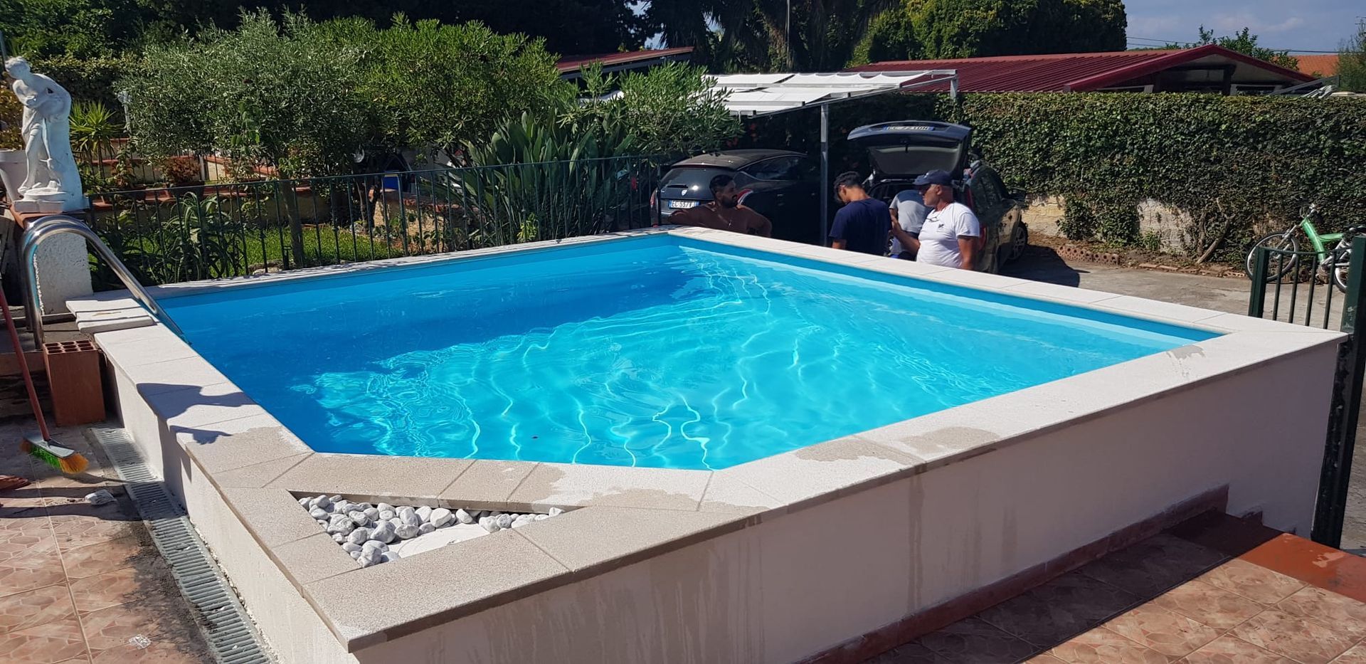 Una piscina rettangolare piena di acqua blu; sullo sfondo due persone vicino a un'auto.