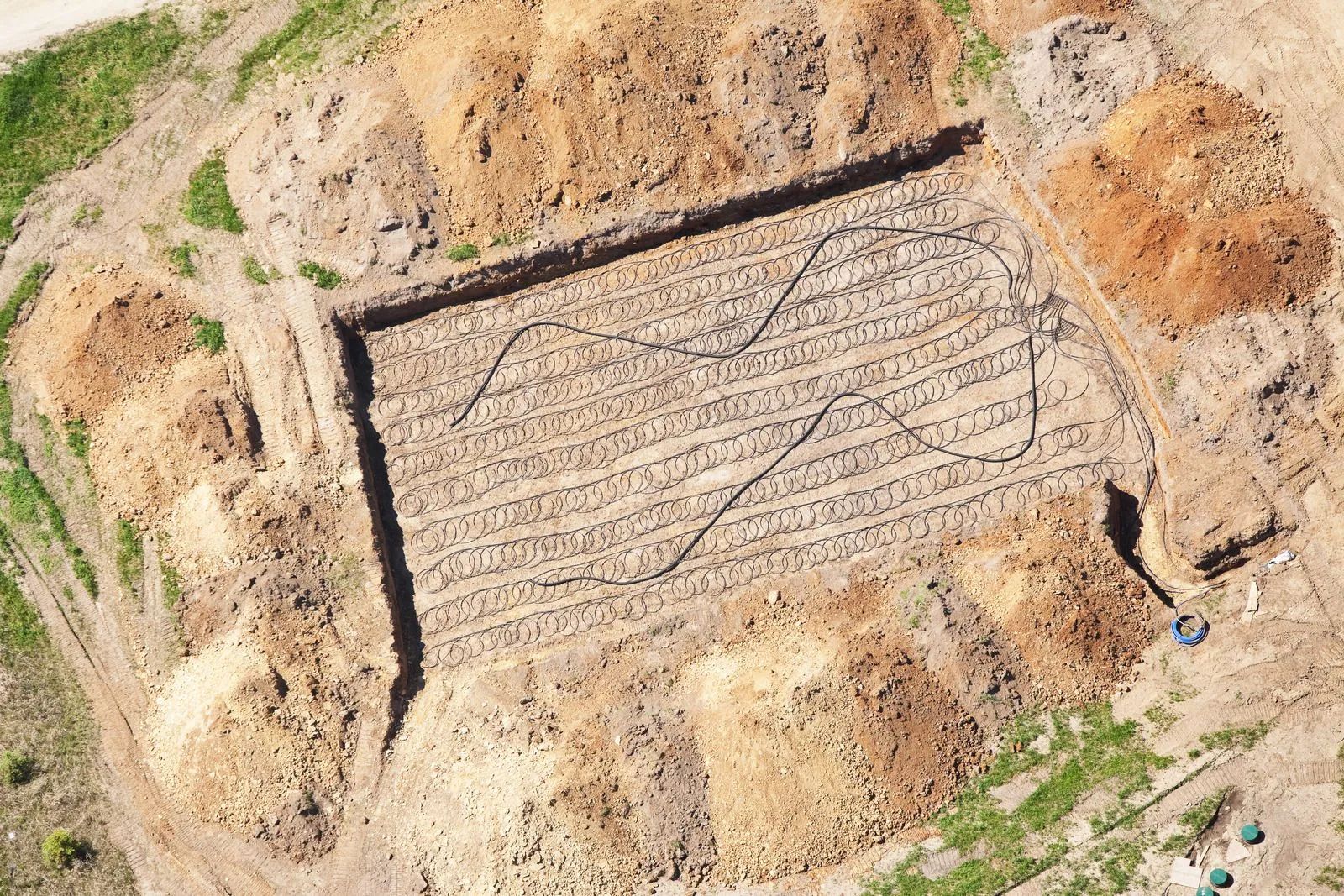 Aerial view of an archaeological dig site; square trench with neat rows, surrounded by soil piles; black line curves across.