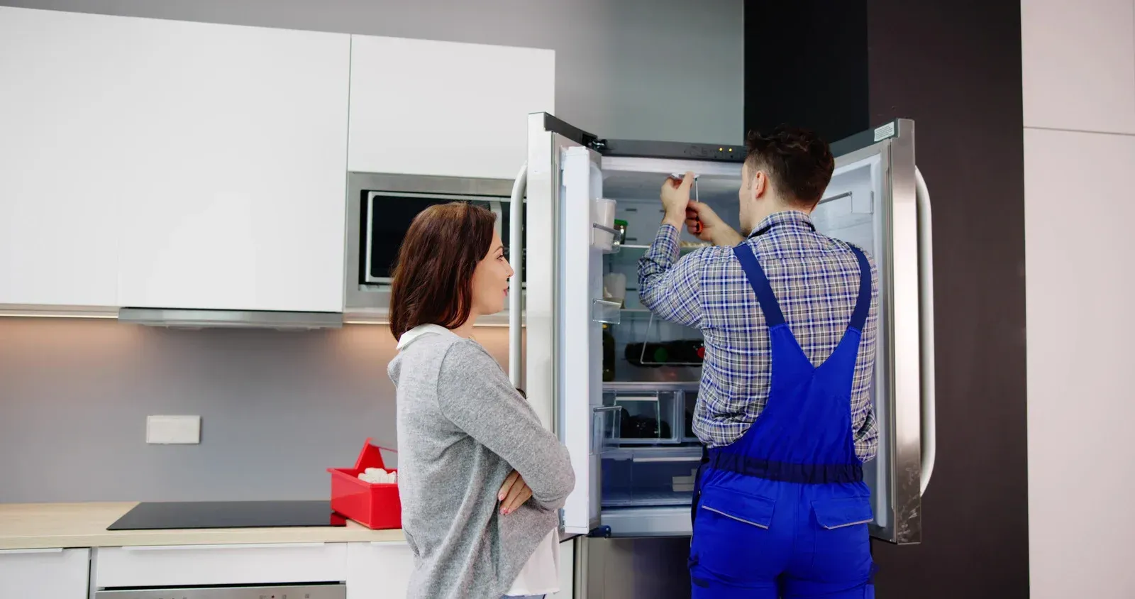 A repairman fixes a refrigerator while a woman watches in a kitchen with white cabinets.