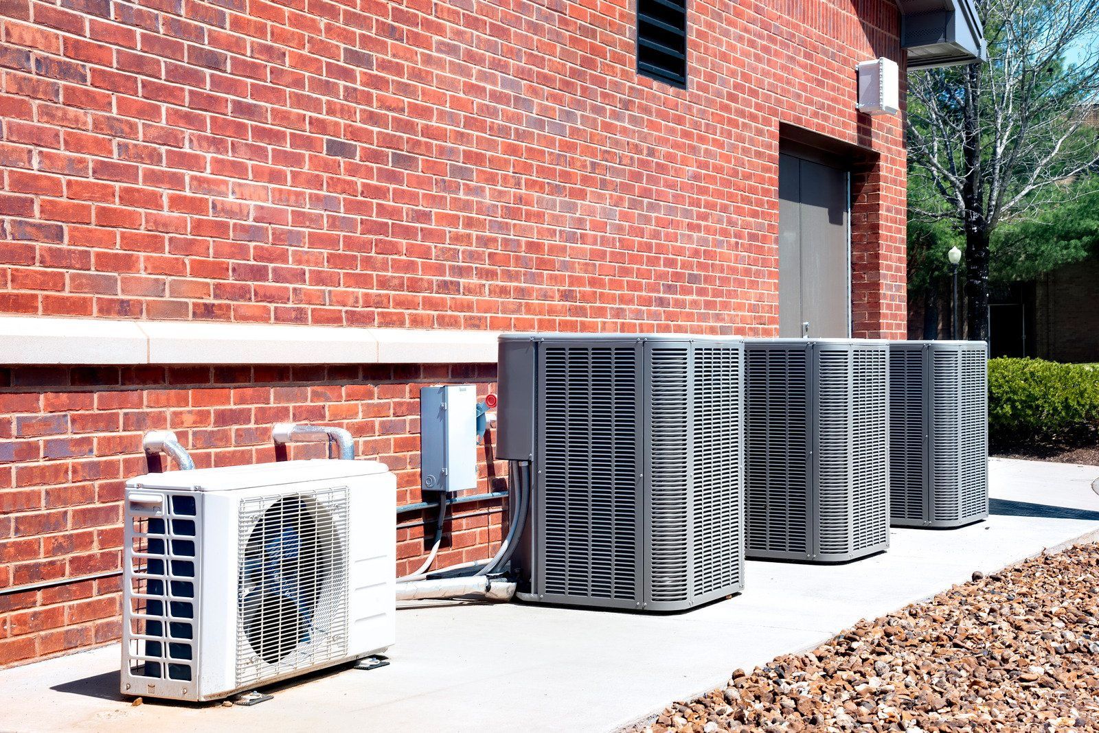 Four air conditioning units are lined up against a brick exterior wall on a concrete pad with rocky ground in the foreground.