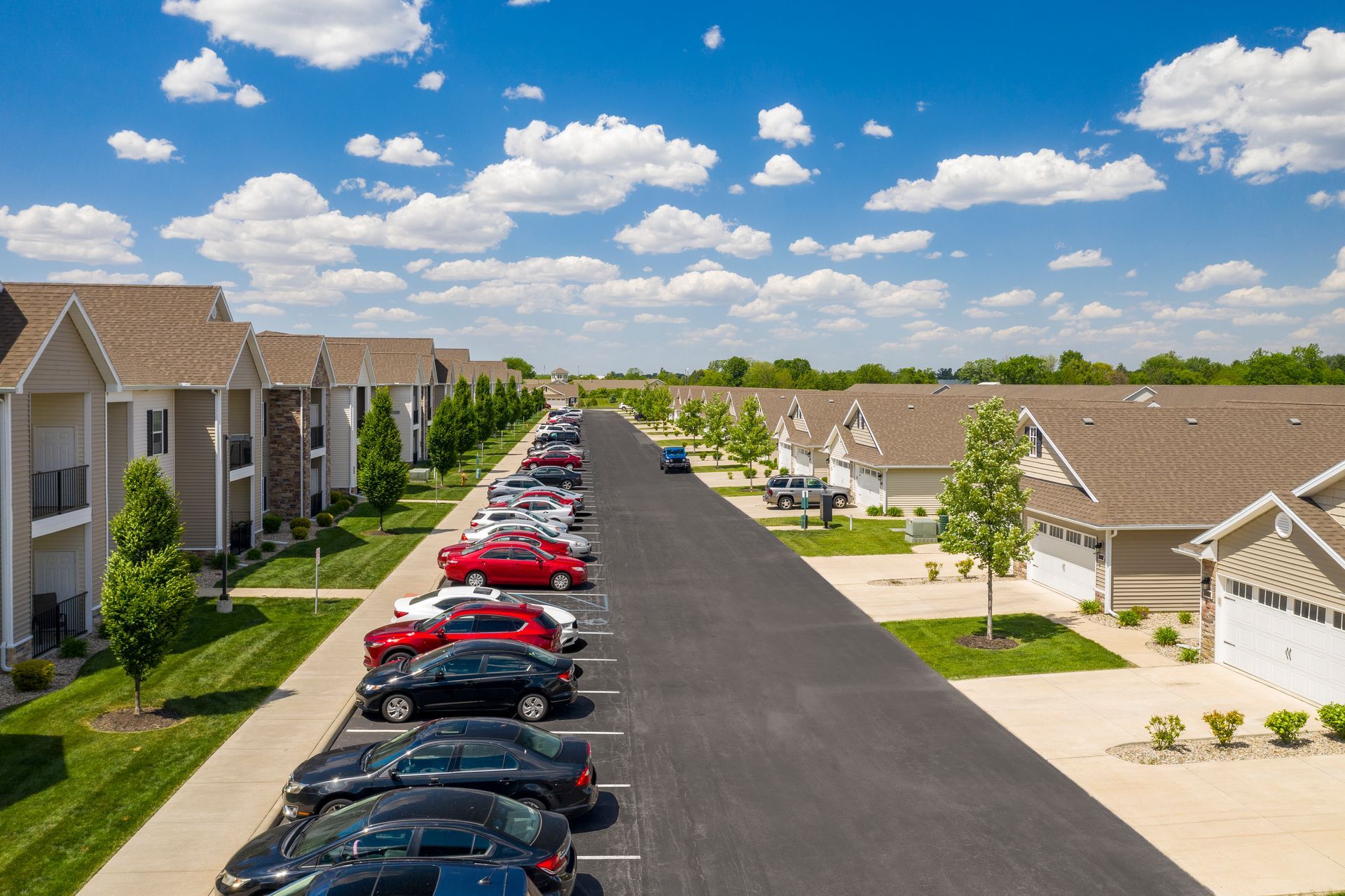 Apartment complex with parked cars along a central road under a blue sky with clouds.