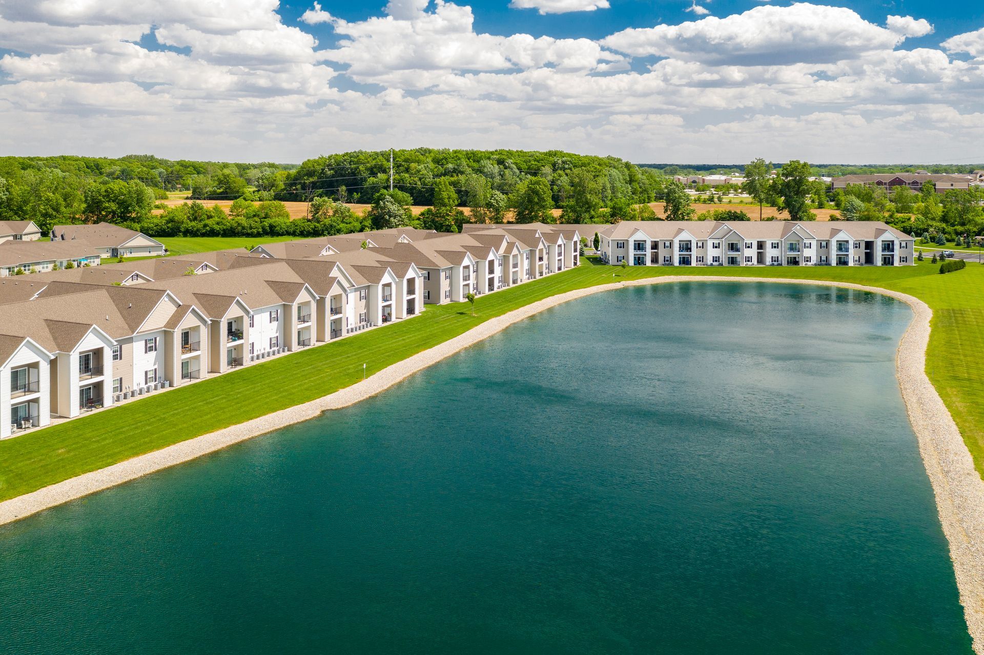 Apartment complex next to a blue lake, with green grass and trees under a cloudy sky.