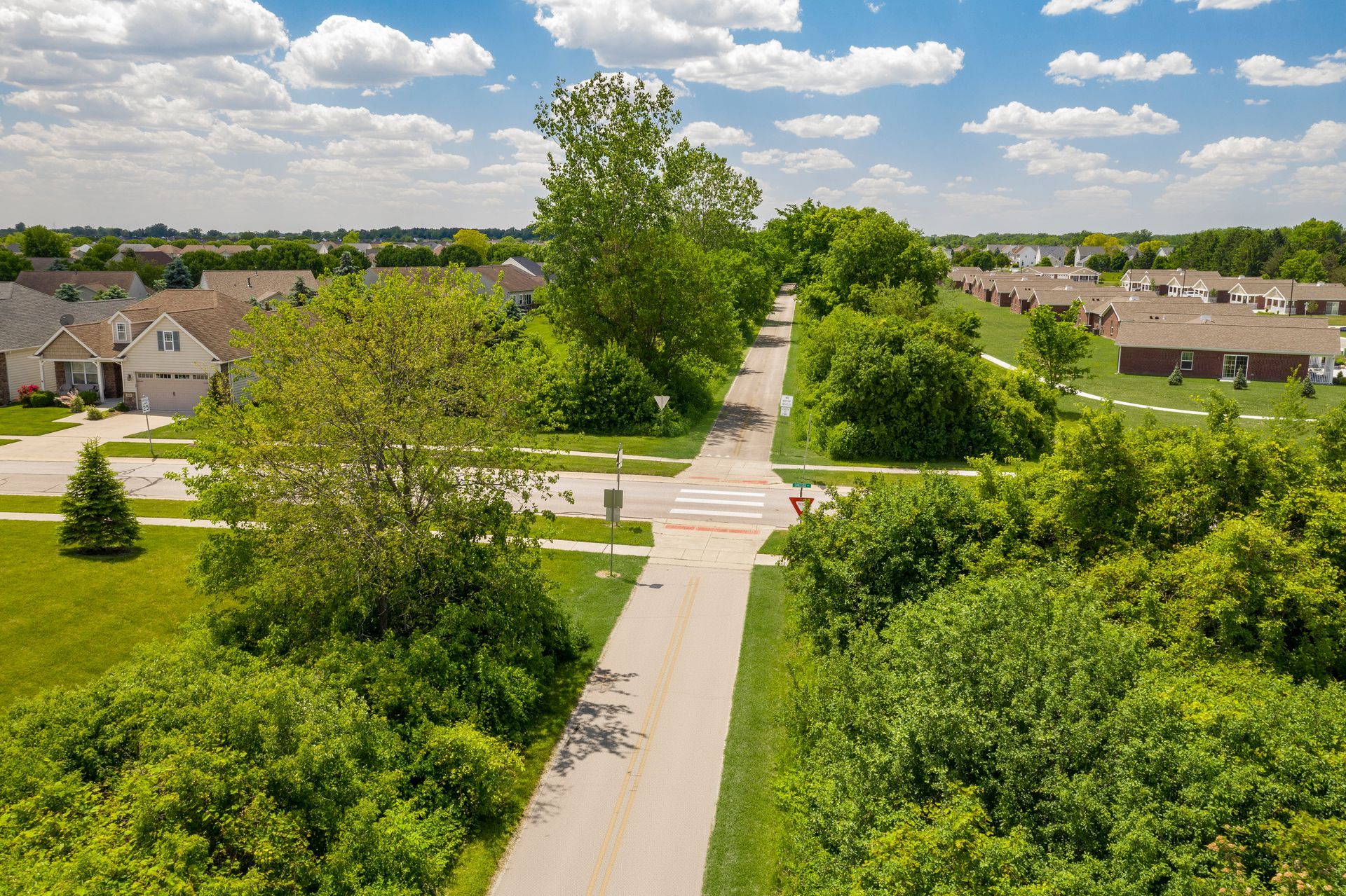 Aerial view of a paved pathway through a residential area with lush green trees and houses under a partly cloudy sky.