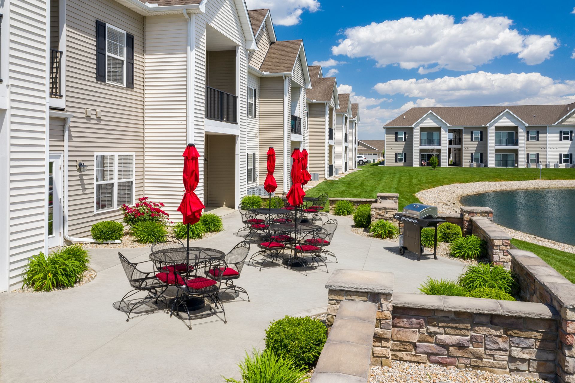 Outdoor patio with red umbrellas, tables, and chairs near a pond and apartment buildings.