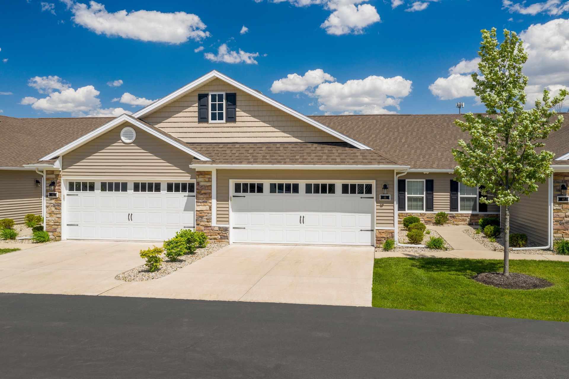 Tan townhomes with white garage doors, blue sky, and green lawn.