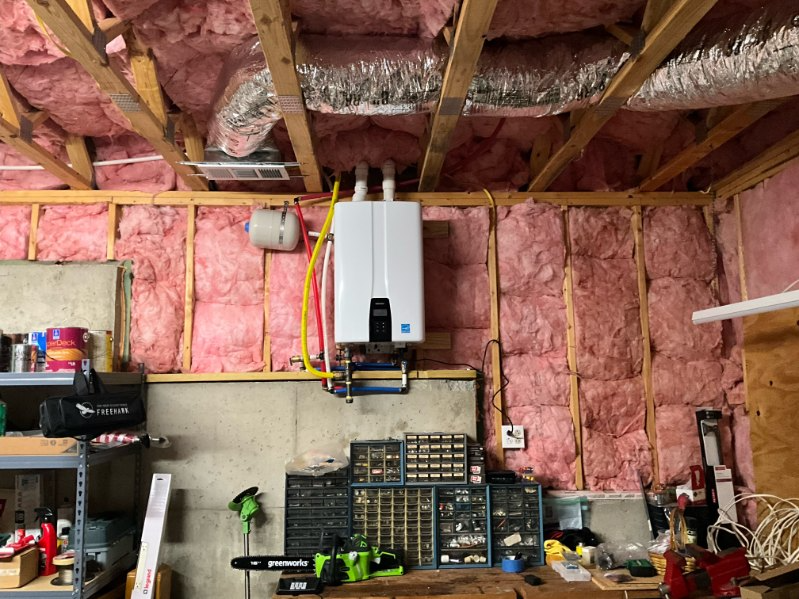 A wall-mounted, white tankless water heater in a garage, surrounded by pink insulation, with tools and storage.
