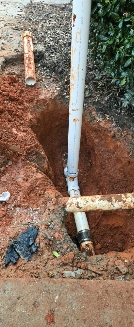 Excavated area with exposed pipes and fittings. Red soil surrounds a white PVC pipe.