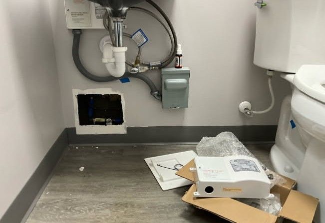 Bathroom with exposed plumbing under a sink and a new water heater in a box on the floor.