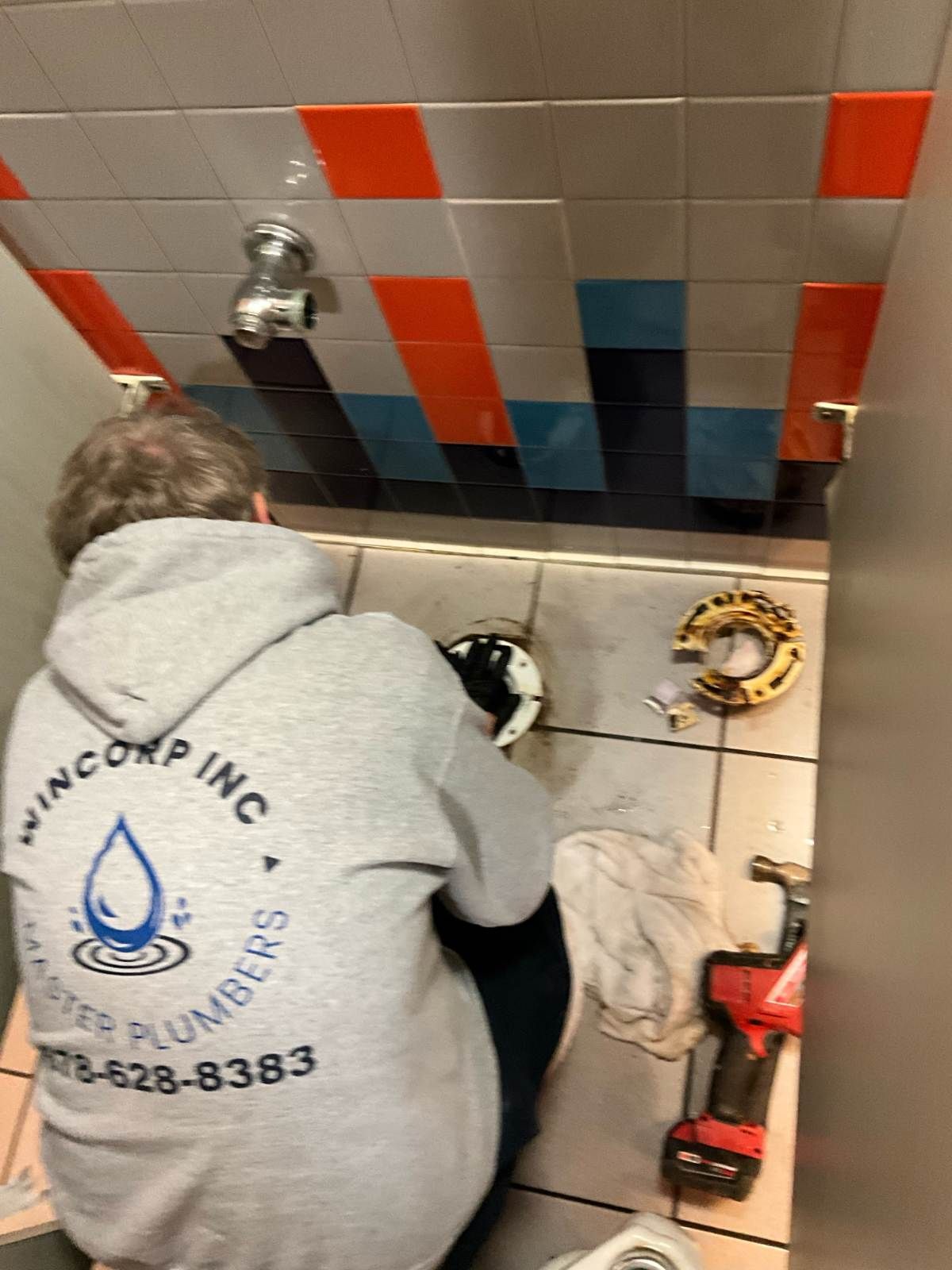 Plumber in a bathroom repairing a toilet. He wears a gray sweatshirt and is surrounded by tools and parts.