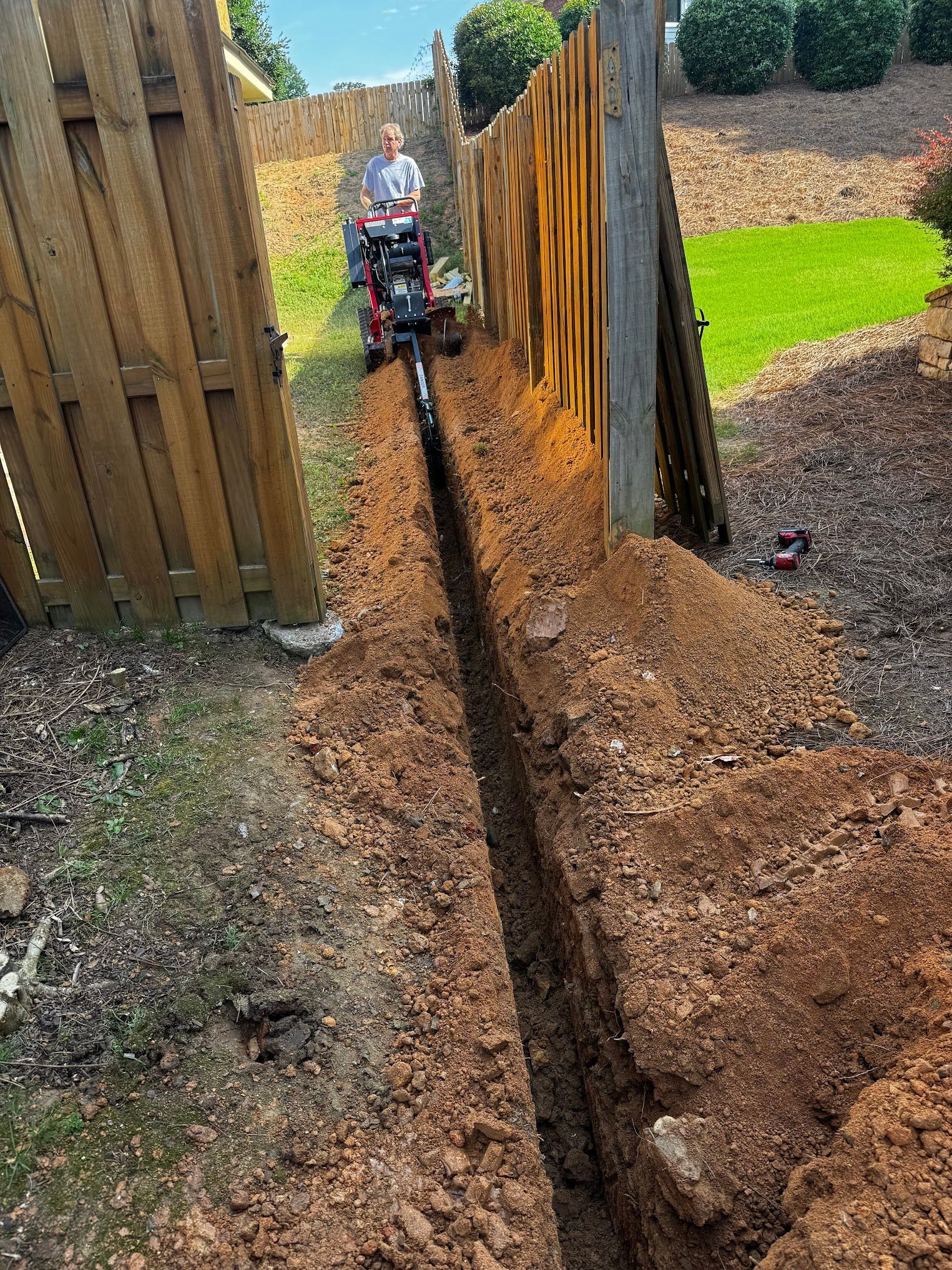 A man is digging a trench in the dirt next to a wooden fence.