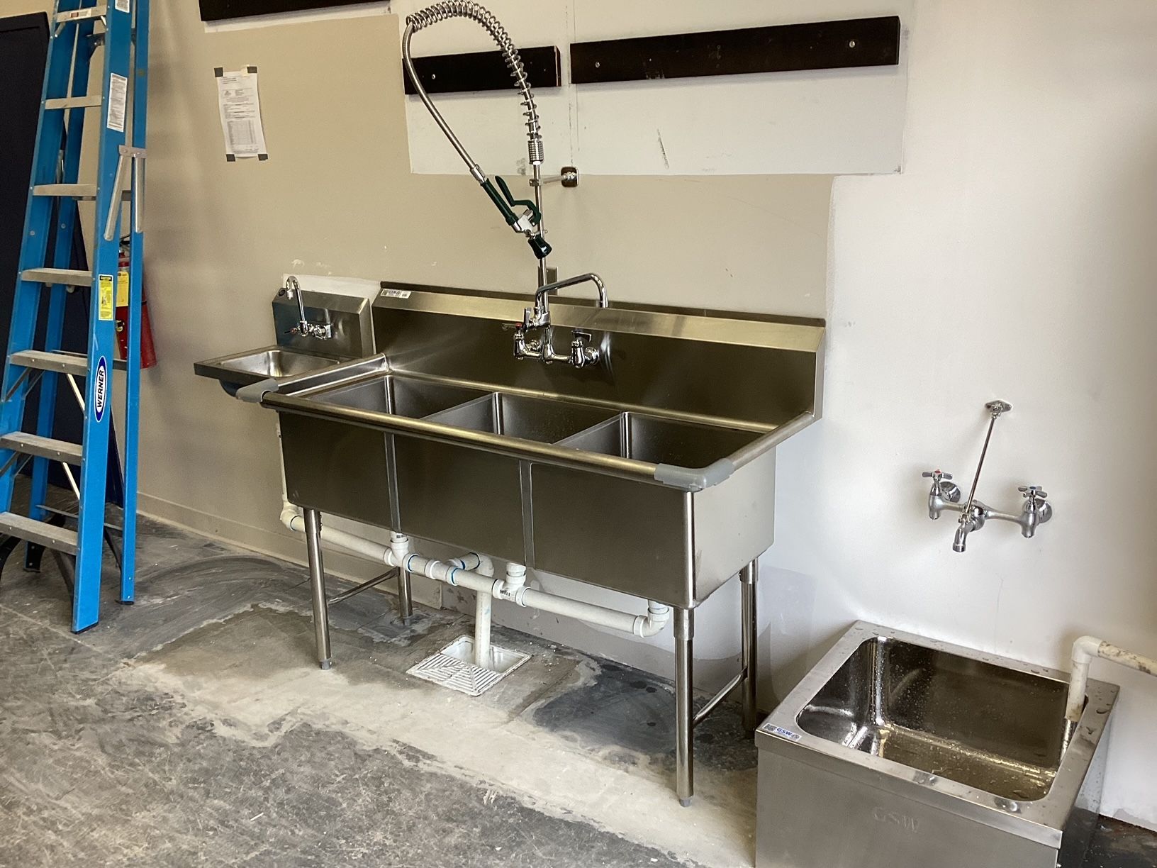 A stainless steel kitchen sink with three sinks and a ladder in the background.
