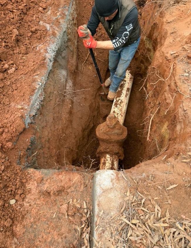 Person digging around a large, rusted pipe in a narrow trench.