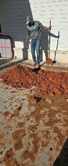 A person wearing a hooded sweatshirt and jeans stands outdoors next to a pile of dirt, holding a shovel.