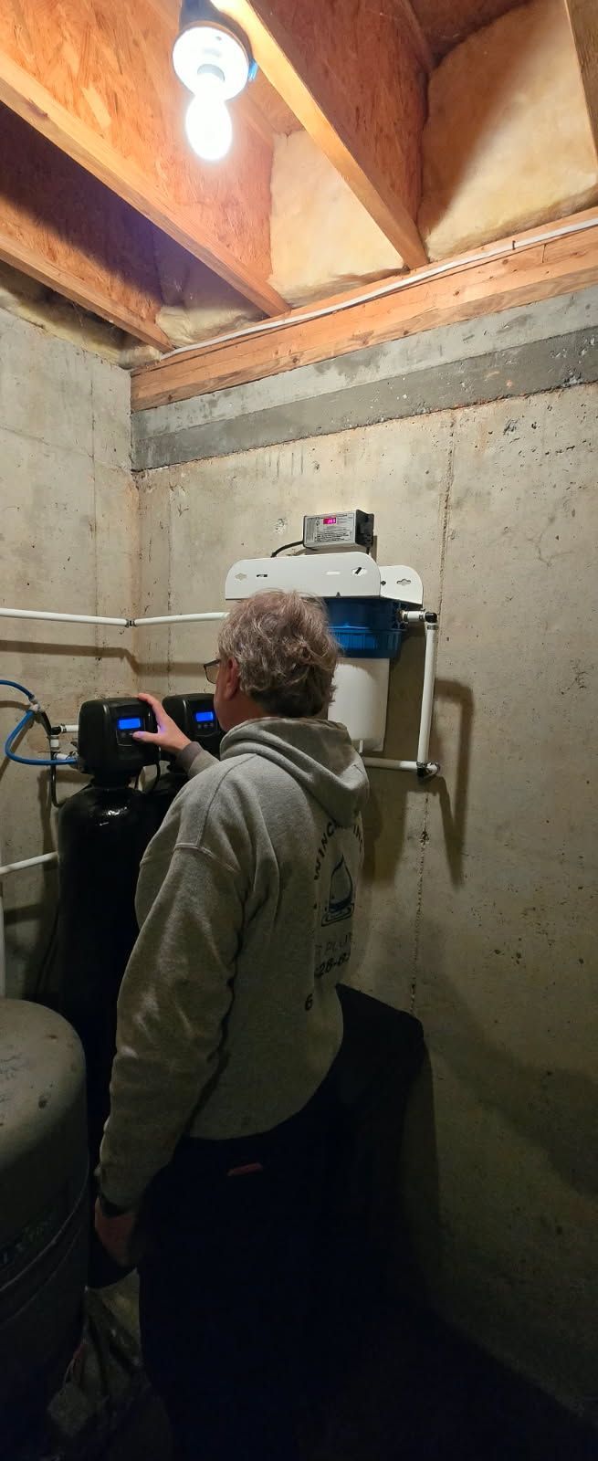 A person adjusting a water filtration system mounted on a concrete basement wall under an exposed wooden ceiling.