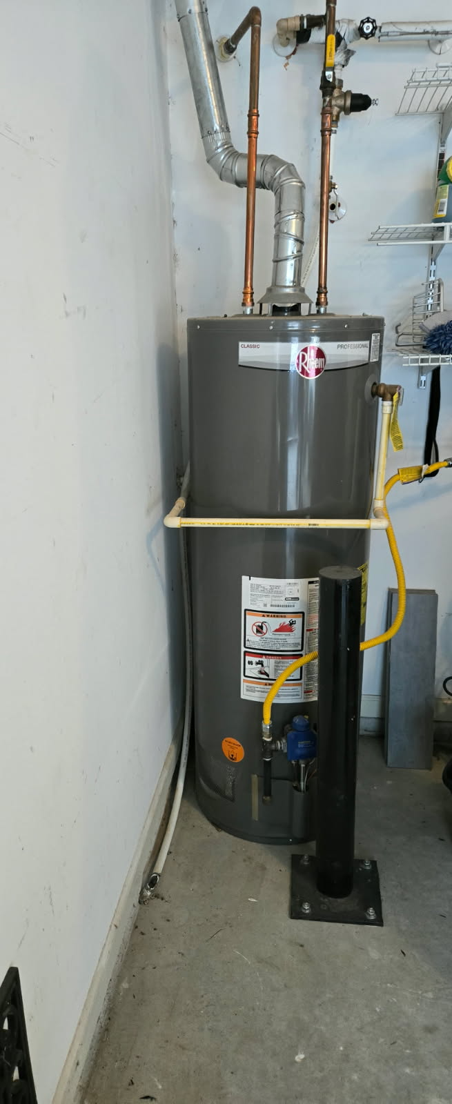 A tall, gray water heater in a garage, with copper pipes and a metal vent. The gas line is yellow.