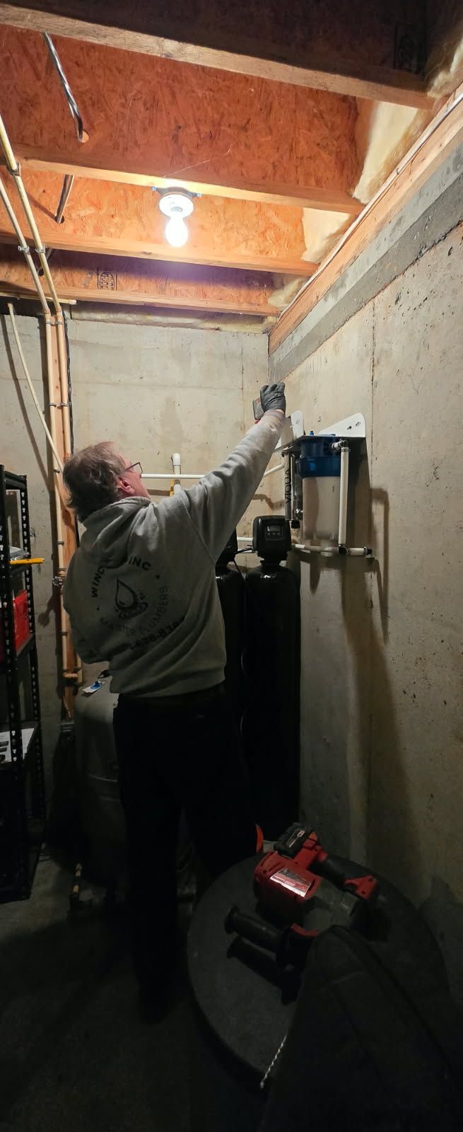 A person in a gray hoodie works on a wall-mounted water filtration system in a utility room with concrete walls.