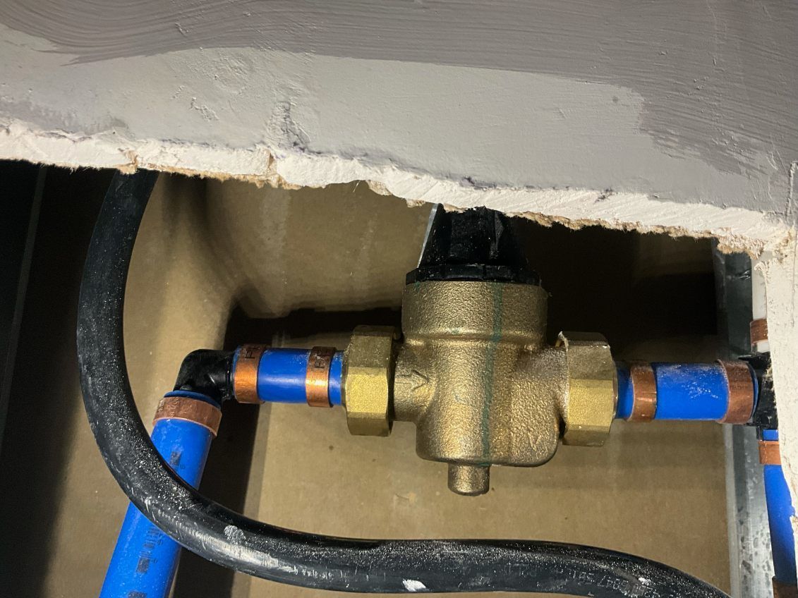 Brass water pressure regulator with blue pipes and black tubing, recessed in a wall opening.