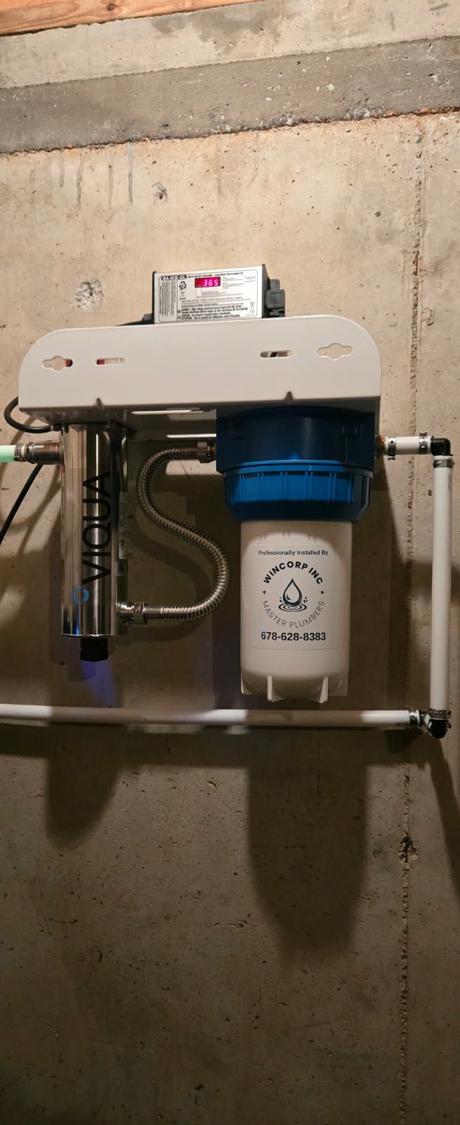 A wall-mounted water filtration system with a stainless steel UV chamber and a white filter housing on a concrete wall.